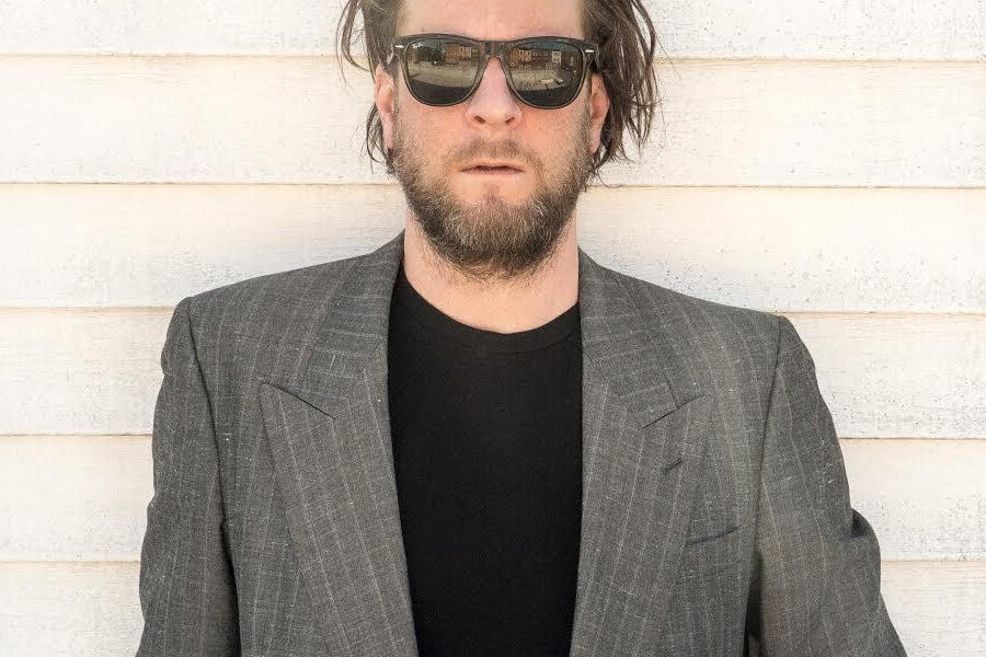 Portrait of Bryce Janssens wearing sunglasses and a dark jacket against a light wall