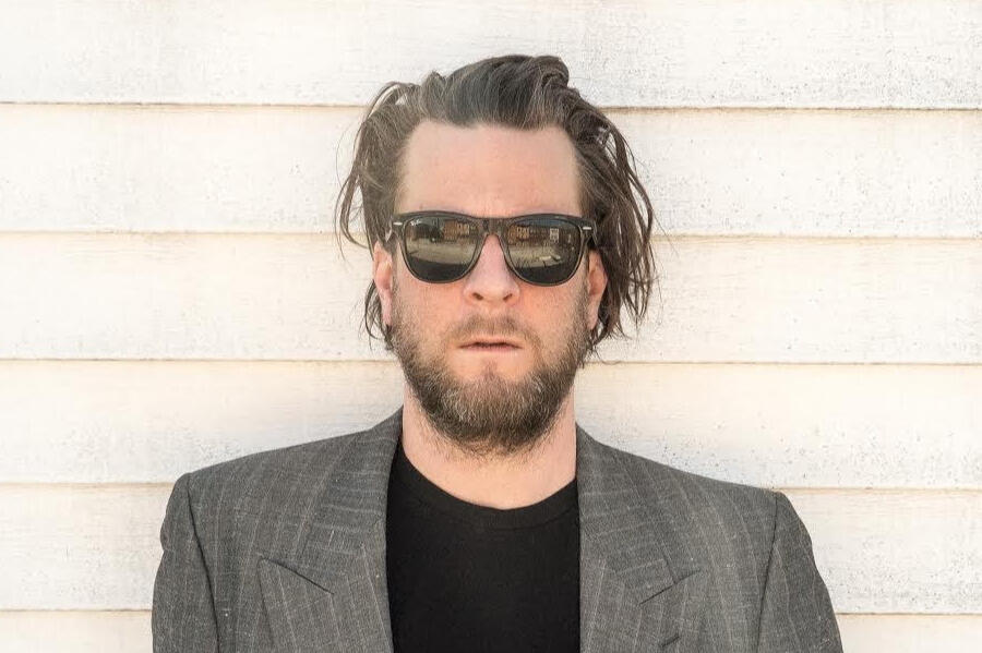 Portrait of Bryce Janssens wearing sunglasses and a dark jacket against a light wall